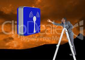 Businessman on a Ladder trying to hold a blue icon against an orange mountain background