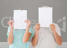 Man and woman covering their face behind paper against grey background