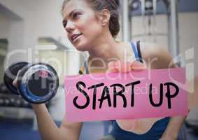Hand holding placard with text start up and woman exercising with dumbbell in background