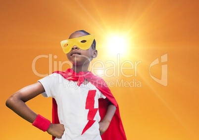 Super kid in red cape and yellow mask standing with hand on hip against bright sunlight