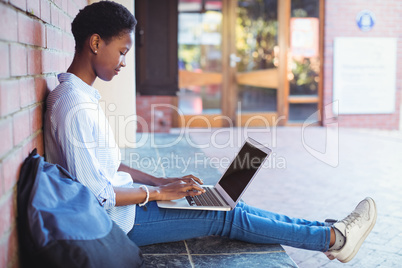 Attentive schoolgirl sitting against brick wall and using laptop
