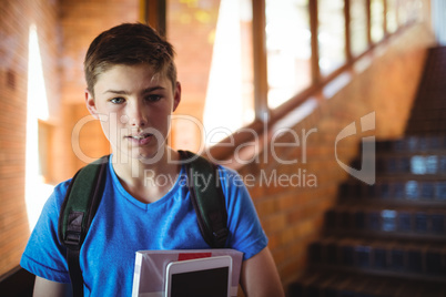 Portrait of schoolboy holding digital tablet and book near staircase