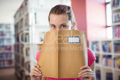 Portrait of schoolgirl hiding her face with book in library