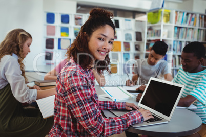 Smiling schoolgirl using laptop with her classmates studying in background