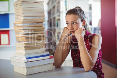 Sad schoolgirl looking at stack of books in library