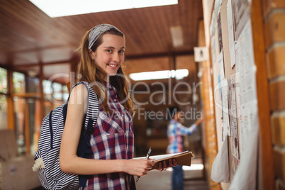 Portrait of smiling schoolgirl standing with book near notice board in corridor