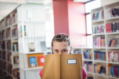 Portrait of schoolgirl hiding her face with book in library