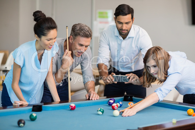 Business executive taking picture with mobile phone while colleagues playing pool