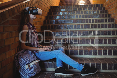 Schoolgirl using virtual reality headset and laptop on staircase