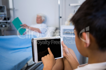 Boy using digital tablet while senior patient reading a book in the background