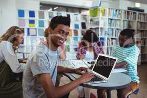 Smiling schoolboy using laptop with his classmates studying in background