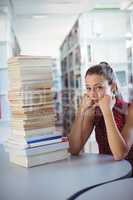 Sad schoolgirl looking at stack of books in library