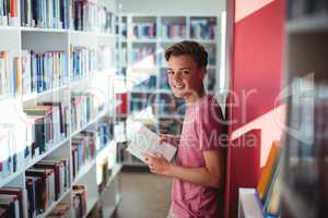 Portrait of happy schoolboy reading book in library