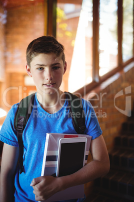 Portrait of schoolboy holding digital tablet and book near staircase