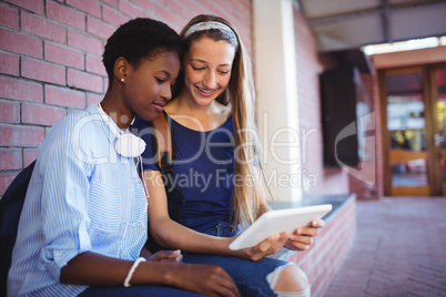 Schoolgirls sitting against brick wall and using digital tablet