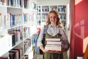 Portrait of schoolgirl standing with stack of books in library