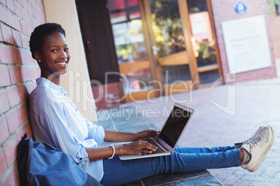 Portrait of happy schoolgirl sitting against brick wall and using laptop