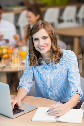 Smiling businesswoman writing on notepad while using laptop in office cafeteria