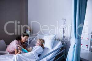 Girl on a hospital bed reading book with her mother