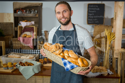 Smiling male staff holding wicker basket of various breads at counter