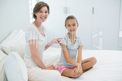 Smiling mother combing daughters hair on bed in bedroom at home