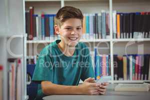 Smiling schoolboy using mobile phone in library at school