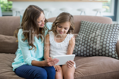 Smiling mother and daughter sitting on sofa using digital tablet in living room