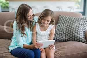 Smiling mother and daughter sitting on sofa using digital tablet in living room