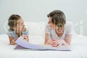 Mother and daughter lying on bed and reading book in bedroom at home