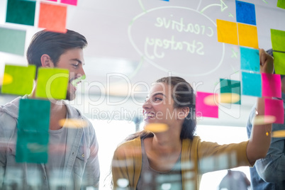 Smiling executives interacting with each other while writing on sticky notes on glass wall