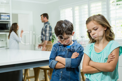 Sad siblings standing with arms crossed while parents arguing in background
