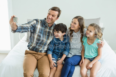 Smiling man taking selfie with family while sitting in bedroom