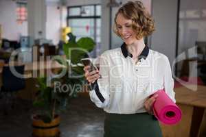 Female graphic designer using mobile phone while holding yoga mat