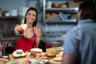Smiling female staff giving heart shape cookie to customer at counter