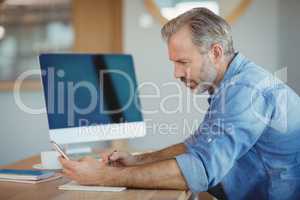 Male business executive writing in organizer while using mobile phone