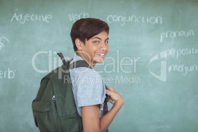 Portrait of schoolboy with backpack standing against chalkboard in classroom