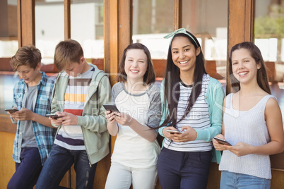 Portrait of smiling school friends using mobile phone in corridor