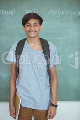 Smiling schoolboy with backpack and book standing against chalkboard in classroom