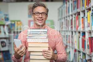 Happy school teacher holding stack of books while using mobile phone in library