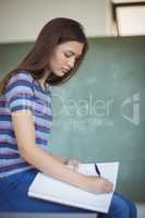 Portrait of schoolgirl sitting on bench and writing on book in classroom