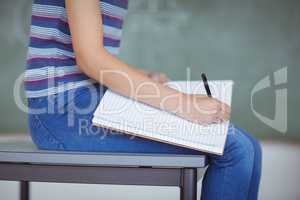Mid section of schoolgirl sitting on bench and writing on book in classroom