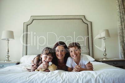 Mother and kids lying on bed with teddy bear in bedroom