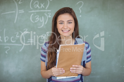 Portrait of schoolgirl holding books against chalkboard in classroom