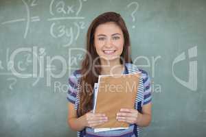 Portrait of schoolgirl holding books against chalkboard in classroom