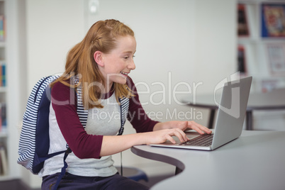 Happy schoolgirl with schoolbag using laptop in library