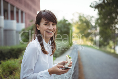Smiling executive having veg roll while using mobile phone on road