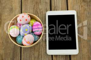 Painted Easter eggs in a bowl and digital tablet on wooden surface