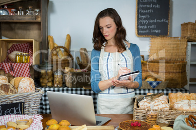 Staff looking at laptop while writing in diary at bakery counter