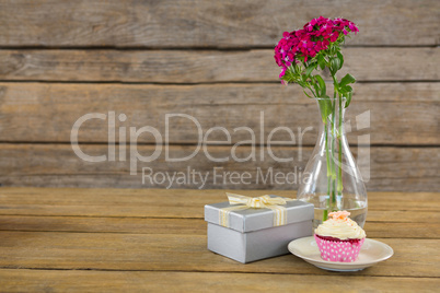 Gift box and flower vase with cupcake in plate on wooden plank