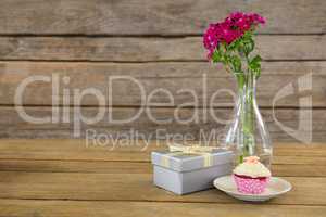 Gift box and flower vase with cupcake in plate on wooden plank